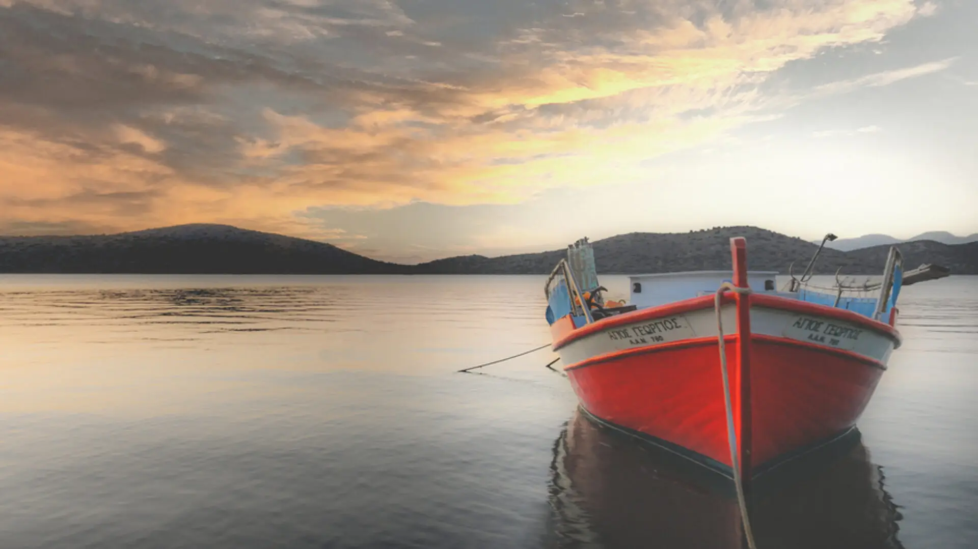 A vibrant red and white fishing boat named "AGIOS GEORGIOS" floats peacefully on calm, reflective water under a dramatic, cloudy sunset sky with distant hills.